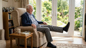 A man sat in a chair with one leg extended, doing a seated leg lift exercise to strengthen his weak legs.